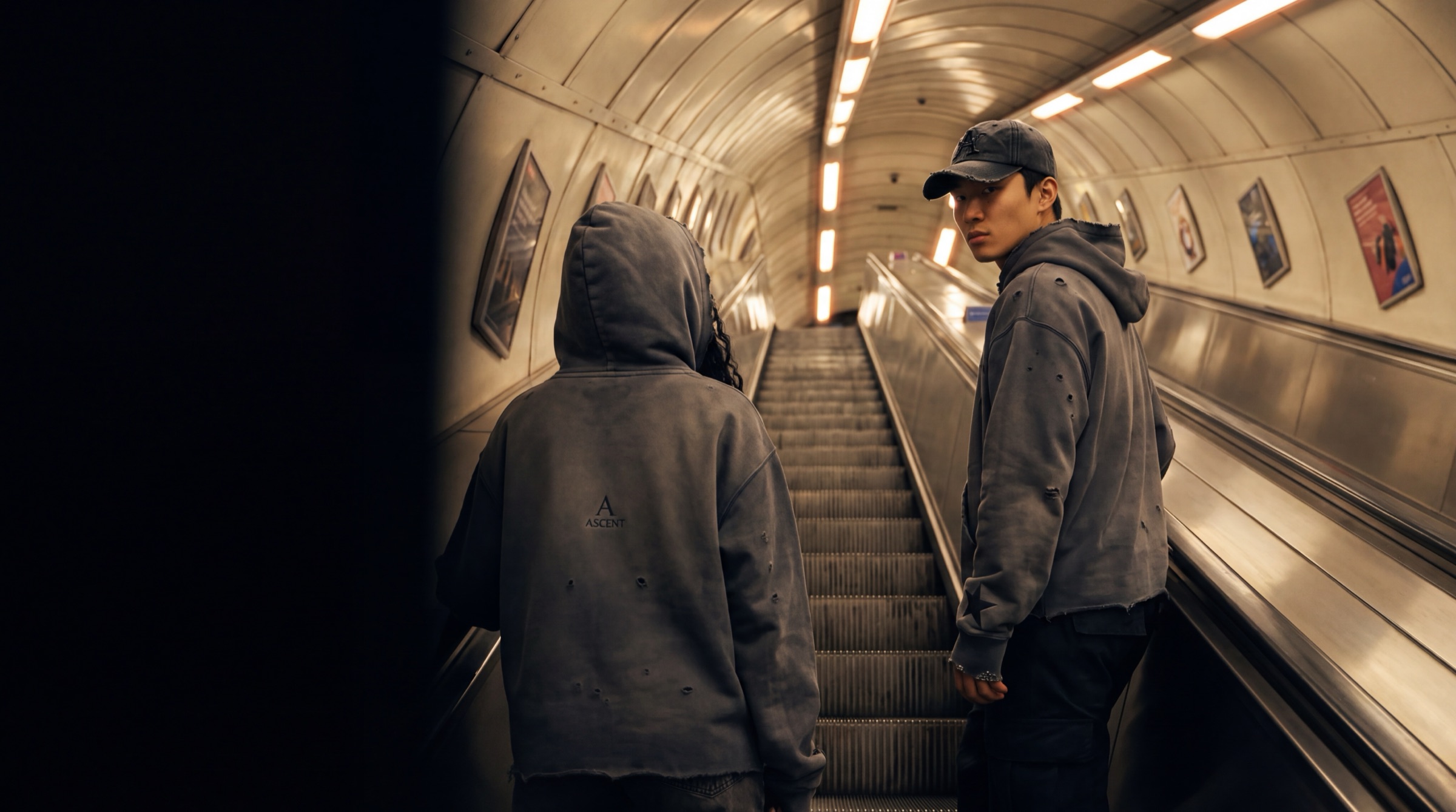 Two hooded figures riding an underground escalator upward at night